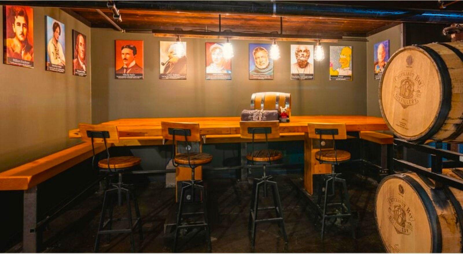 The Distiller’s Table room: a long wooden table with bar stools, framed portraits along the walls and Chuckanut Bay barrels in the corner