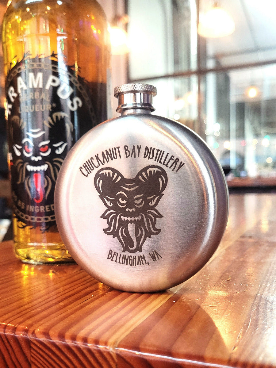 Stainless steel flask with 'Chic Kanut Bay Distillery' logo on a wooden surface with a blurred background.