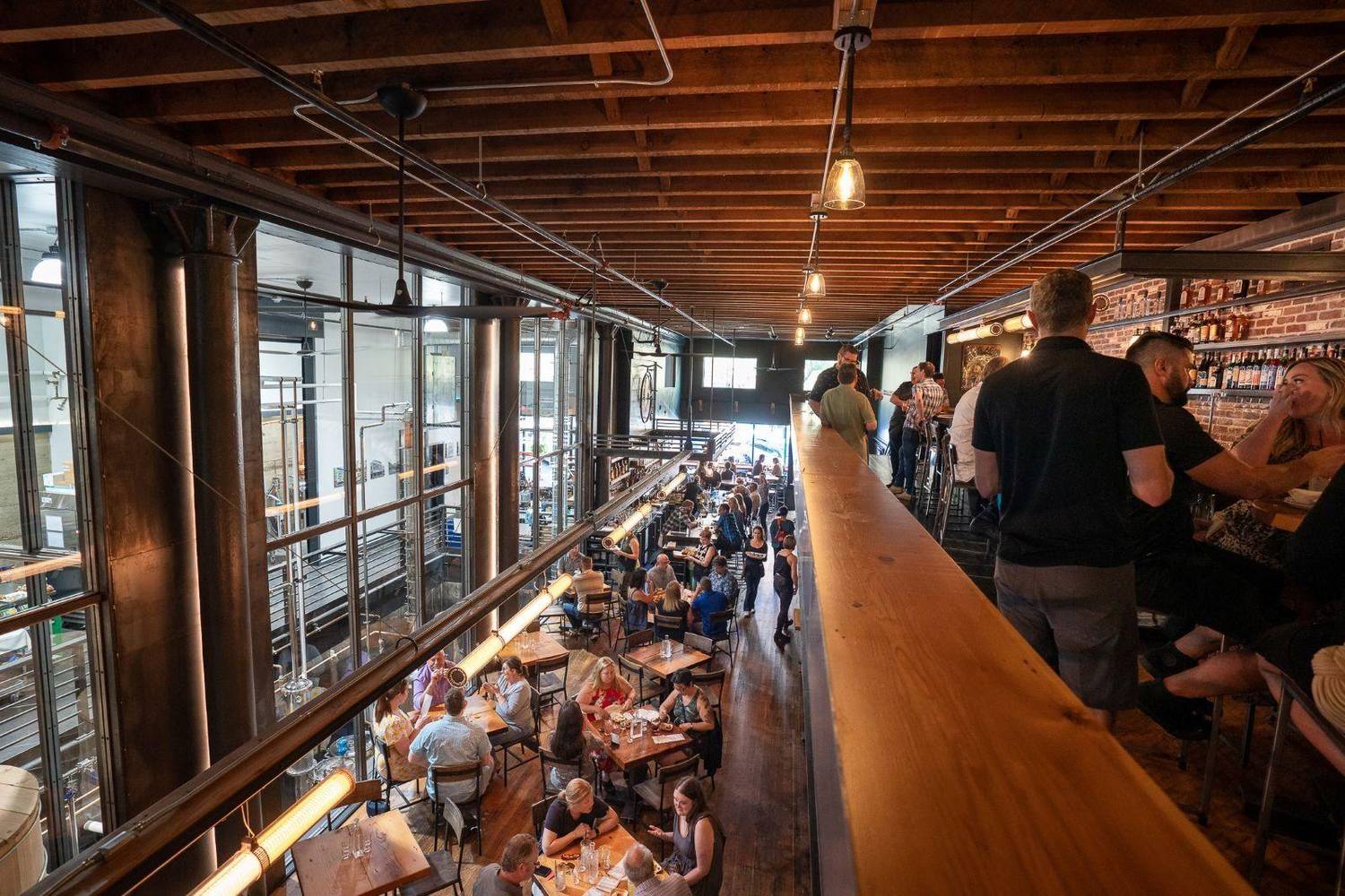 Penny Farthing seen from the mezzanine: guests dining below and gathered at the upstairs bar, with the distillery behind the glass