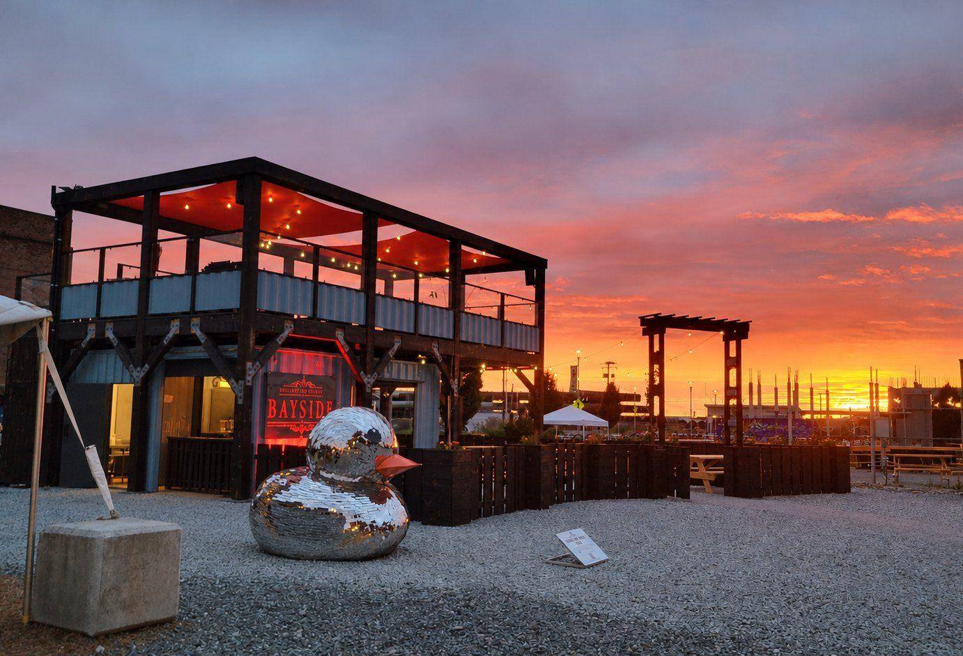Bayside’s container bar at sunset, with the rooftop deck under red sails and the mirrored duck sculpture out front