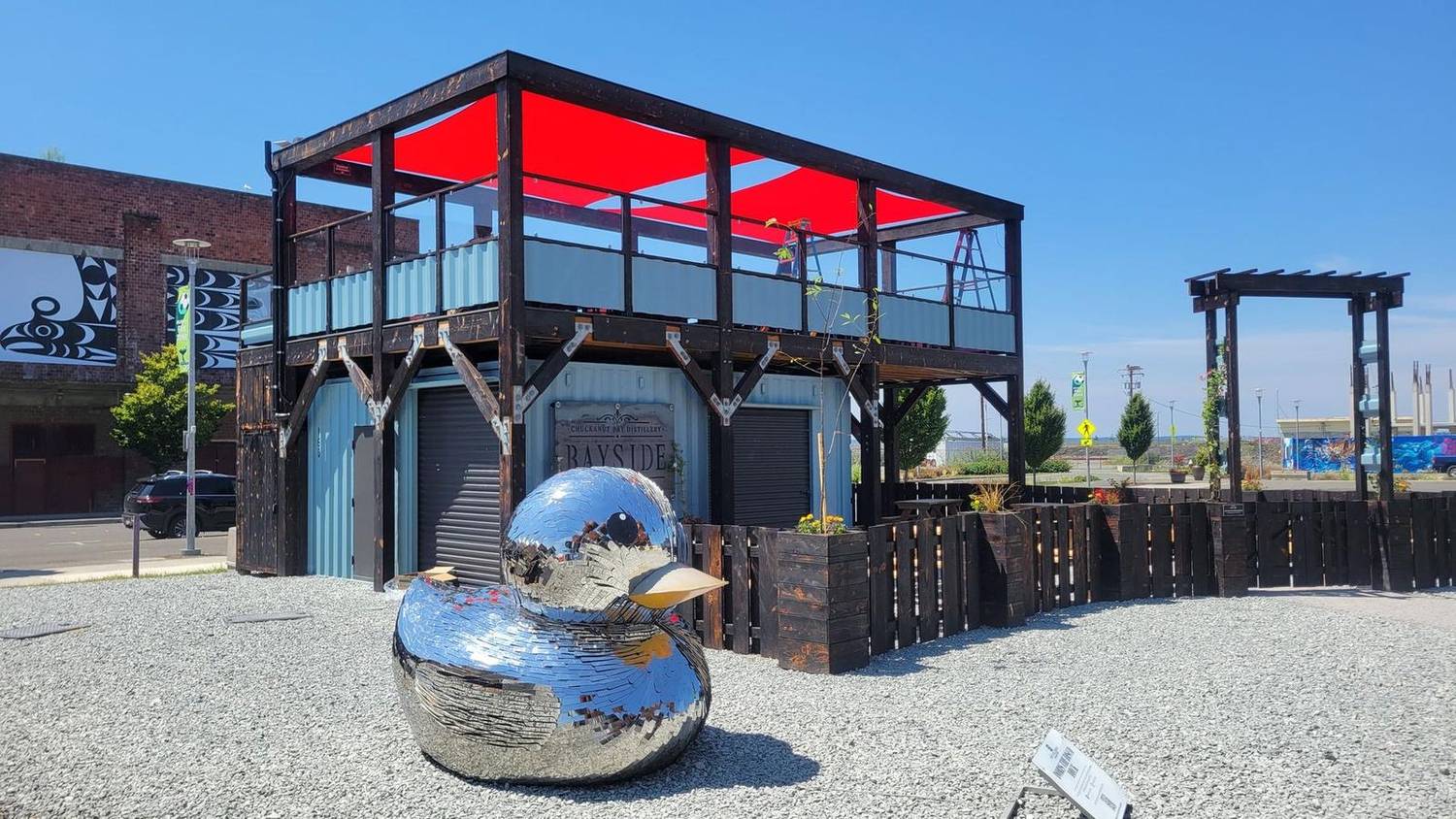 Bayside's two-story container bar with red shade sails on the rooftop deck and a giant mirrored duck sculpture out front